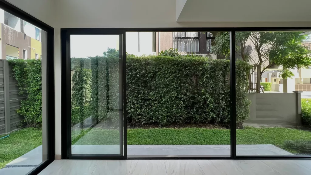 Widok z nowoczesnego wnętrza przez duże przesuwne okna na niewielki ogródek z trawnikiem i gęstym żywopłotem, z drzewami i fragmentami sąsiednich budynków w tle.