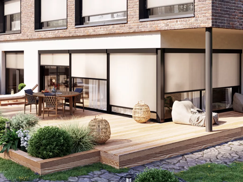 Nowoczesny taras z drewnianym podestem przy domu z dużymi przeszkleniami osłoniętymi roletami zewnętrznymi; stół z krzesłami, leżanka, pufy i wiklinowe lampiony wśród roślin.