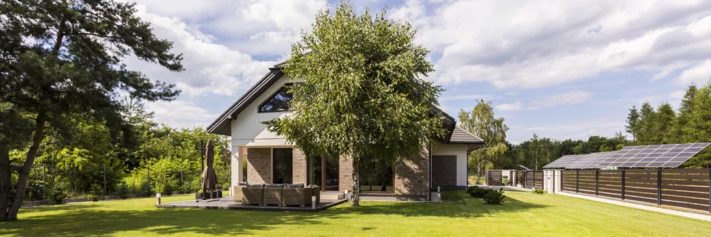 Nowoczesny dom jednorodzinny z dwuspadowym dachem, oknami i drzwiami zewnętrznymi, dużym trawnikiem i tarasem z meblami, częściowo zasłonięty przez drzewo; przy ogrodzeniu widoczne panele fotowoltaiczne.