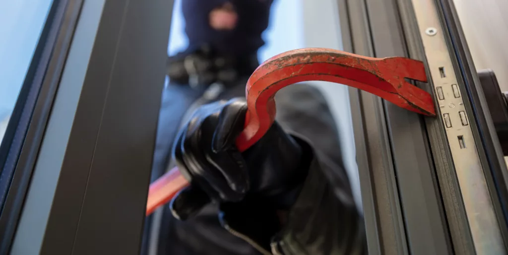 Close-up of a person wearing a balaclava and black gloves, prying open an aluminum window with a red crowbar.