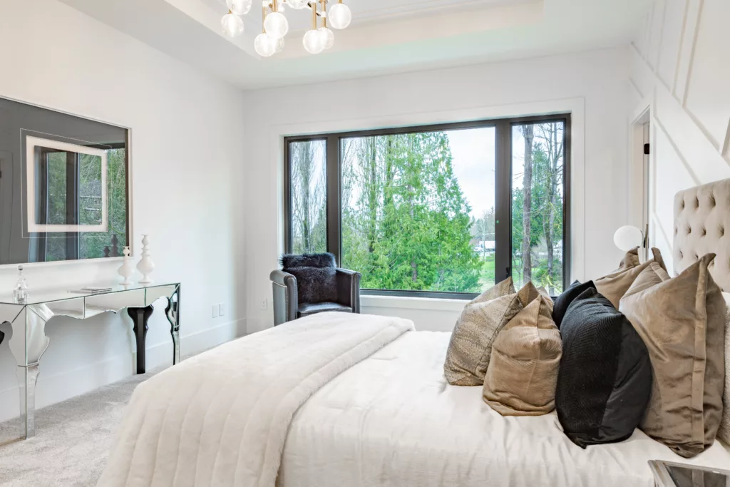 Jasna, nowoczesna sypialnia z dużym oknem na zieleń, łóżkiem z beżowym pikowanym wezgłowiem i wieloma poduszkami, lustrzaną konsolą oraz fotelem przy oknie.