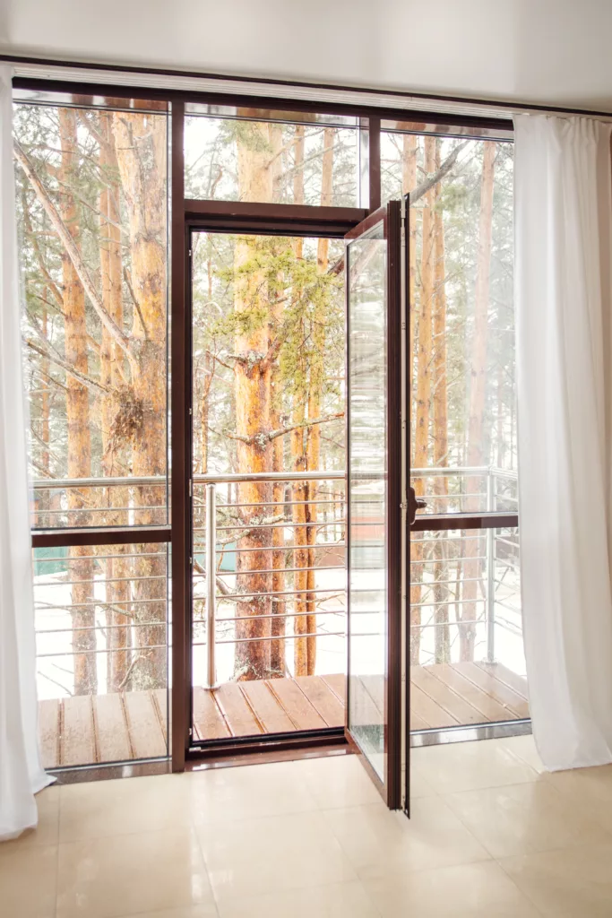 Otwarte przeszklone drzwi balkonowe z białymi zasłonami prowadzą na drewniany balkon z metalową balustradą, z widokiem na zaśnieżony las sosnowy.