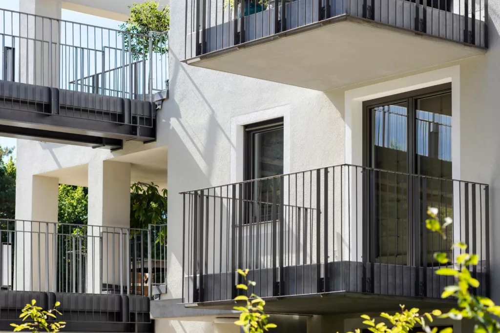 Fasada nowoczesnego bloku z kilkoma balkonami o metalowych balustradach z czarnymi oknami, białym tynkiem i roślinnością w tle, oświetlona słońcem.