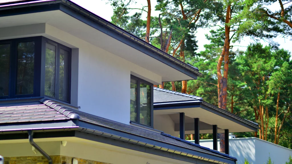 Nowoczesny dom z białą elewacją, dużymi oknami i grafitowym dachem wielospadowym, na tle sosnowego lasu.