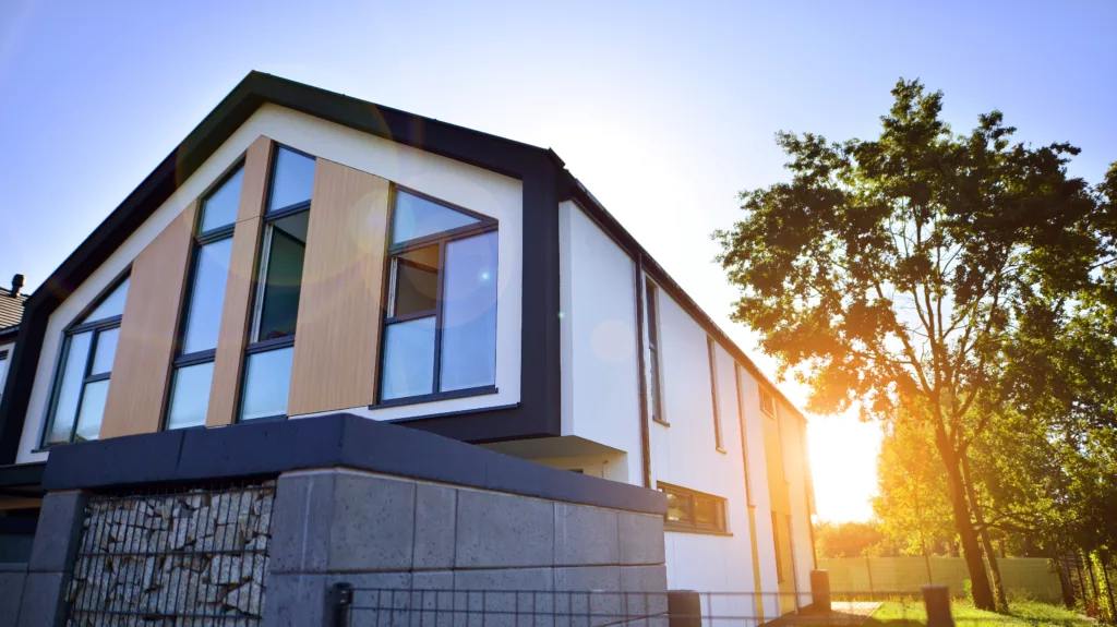 Nowoczesny, dwukondygnacyjny dom z dużymi oknami i panelami drewnianymi na białej elewacji, sfotografowany z dołu w popołudniowym słońcu, obok wysokiego drzewa i betonowego ogrodzenia.