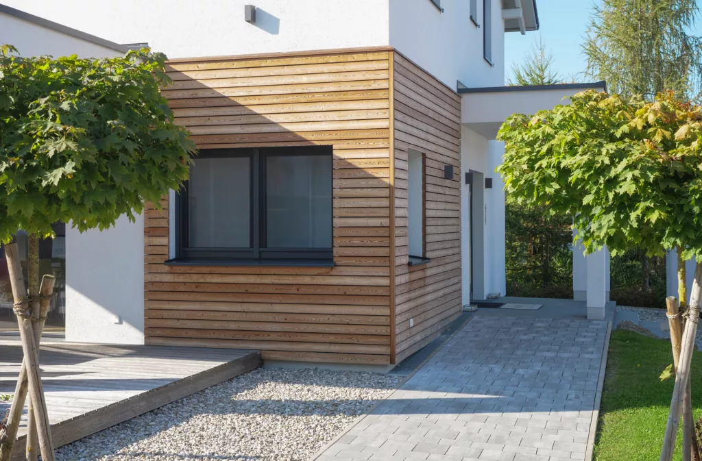 Nowoczesna elewacja domu z poziomą drewnianą okładziną i białym tynkiem, z dużym ciemnym oknem, brukowaną ścieżką, pasem żwiru i zadbanymi drzewami w słoneczny dzień.