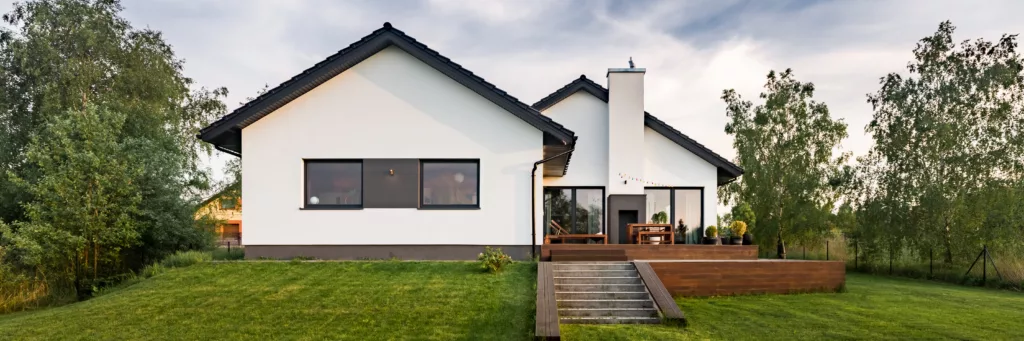 Nowoczesny parterowy dom z białą elewacją i ciemnym dachem, z dużymi oknami oraz drewnianym tarasem i schodami na zadbany trawnik, otoczony drzewami o zmierzchu.