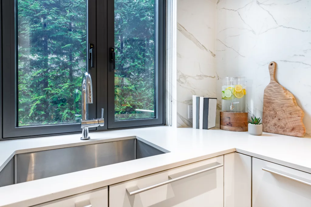 Nowoczesny narożnik kuchenny: biały blat i marmurowa ściana, duży stalowy zlew z chromowaną baterią pod czarnym oknem z widokiem na zieleń, obok dyspenser z wodą i plasterkami cytrusów, sukulent i drewniana deska.