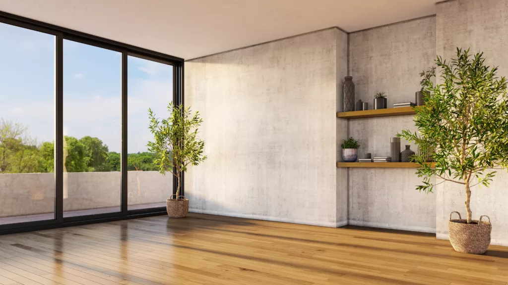 Jasne, minimalistyczne wnętrze z dużymi przeszklonymi drzwiami na taras, drewnianą podłogą, betonową ścianą, półkami z dekoracjami i dwiema roślinami w wiklinowych koszach, z widokiem na zieleń za oknem.
