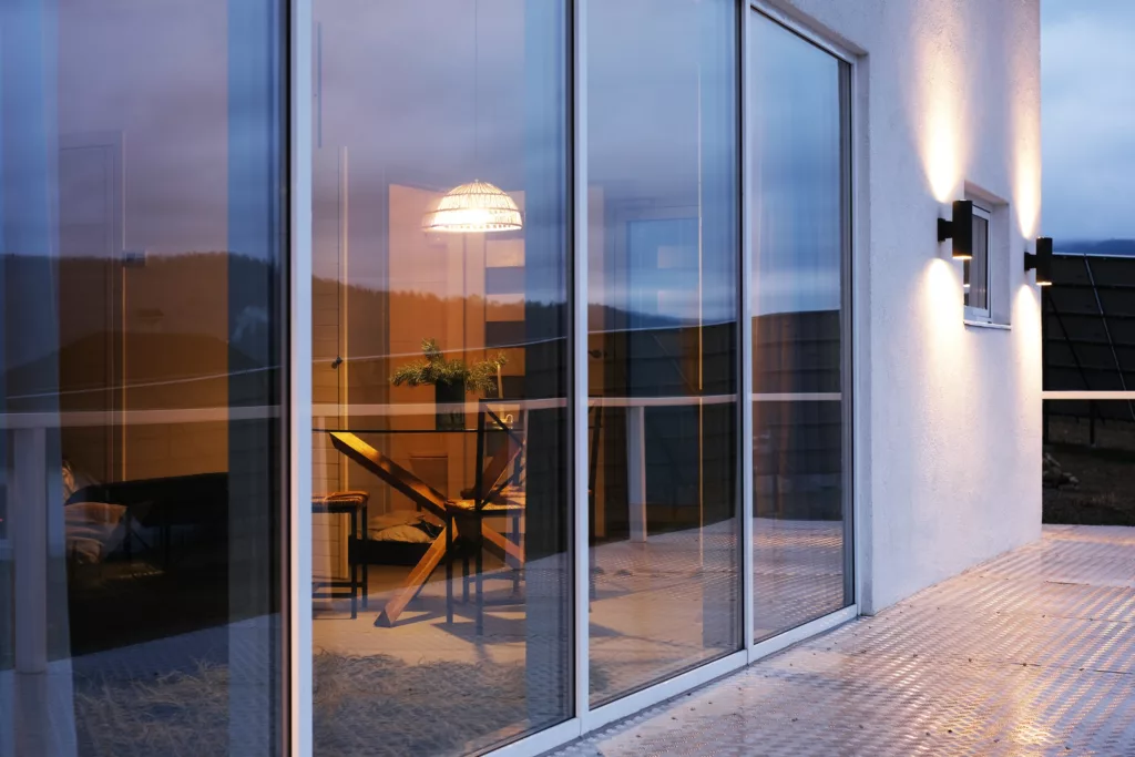 Taras nowoczesnego domu o zmierzchu z dużymi oknami, w którym odbijają się chmury i wzgórza, przez które widać ciepło oświetloną jadalnię z drewnianym stołem i rośliną oraz elewację z dwoma kinkietami rzucającymi światło w górę i w dół.