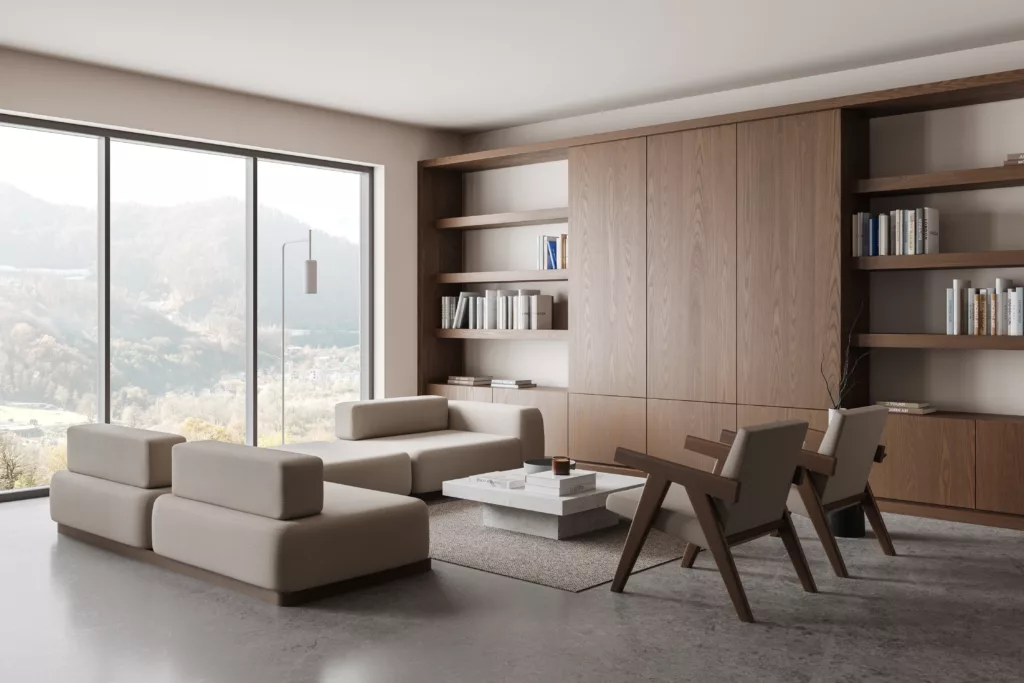 Minimalistyczny salon z dużymi oknami z widokiem na góry, beżowymi modułowymi sofami i dwiema drewnianymi fotelami wokół niskiego kamiennego stolika, na tle zabudowy z drewnianymi półkami i książkami.