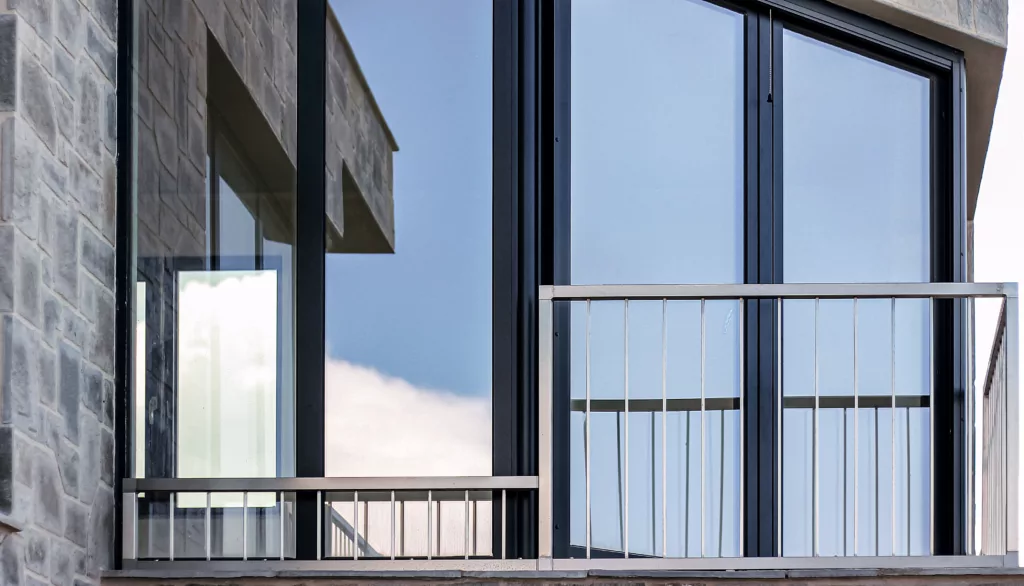 Zbliżenie nowoczesnego balkonu z aluminiową balustradą przy dużych, przeszklonych drzwiach, w których odbijają się niebo i chmury, na tle kamiennej elewacji.