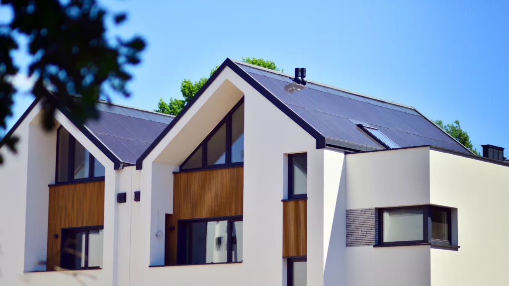 Nowoczesny dom jednorodzinny z białą elewacją i drewnianymi akcentami, dużymi oknami oraz dwuspadowym dachem pokrytym panelami fotowoltaicznymi i świetlikiem, na tle błękitnego nieba i drzew.