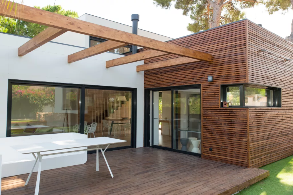 Nowoczesny parterowy dom z płaskim dachem, drewnianą elewacją i dużymi przeszklonymi drzwiami przesuwnymi, z tarasem z desek i pergolą z drewnianych belek, otoczony zielenią.