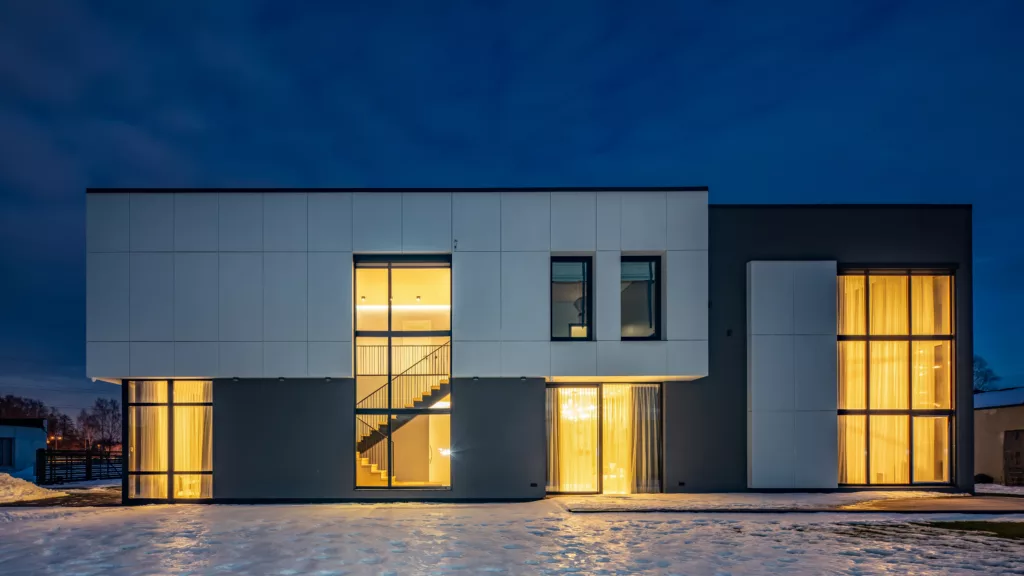 Nowoczesny, dwukondygnacyjny dom z płaskim dachem i dużymi przeszkleniami, oświetlony ciepłym światłem wewnątrz, sfotografowany o zmierzchu na tle ciemnoniebieskiego nieba, ze śniegiem przed budynkiem i widocznymi przez centralne okno schodami.