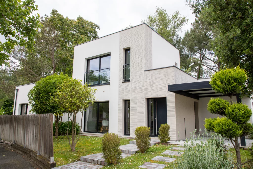Nowoczesny, dwukondygnacyjny dom z płaskim dachem, białą i jasnoszarą elewacją, dużymi oknami z czarnymi ramami oraz zadaszonym podjazdem, otoczony ogrodem z drzewami i kamienną ścieżką.