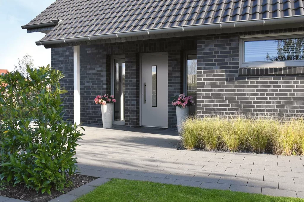 Nowoczesne drzwi wejściowe w jasnoszarym kolorze z wąskim pionowym przeszkleniem i długim czarnym uchwytem, osadzone w elewacji z ciemnej cegły, z wąskimi naświetlami bocznymi i donicami z kwiatami przy brukowanym podeście.
