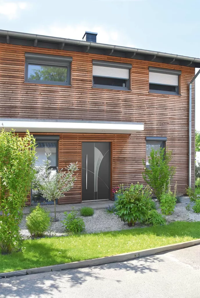 Nowoczesne antracytowe drzwi wejściowe z długim pionowym uchwytem i łukowymi przeszkleniami, osadzone w elewacji z drewnianych desek, otoczone niską zielenią ogrodu frontowego.