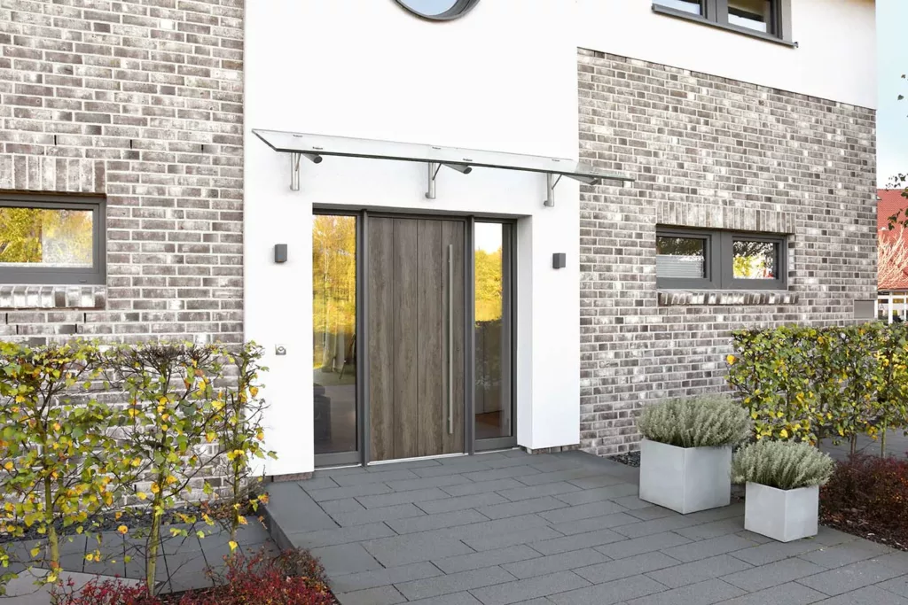 Nowoczesne drzwi zewnętrzne z pionowym uchwytem i wąskimi naświetlami, pod szklanym daszkiem, w elewacji z jasnej cegły, z donicami przy wejściu.
