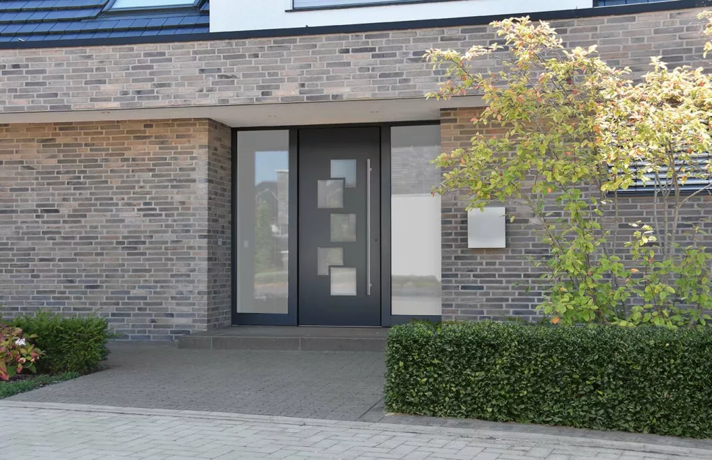 Nowoczesne, grafitowe drzwi wejściowe z czterema kwadratowymi przeszkleniami i wysokimi naświetlami po obu stronach, osadzone w elewacji z szarej cegły przy podjeździe z żywopłotem.