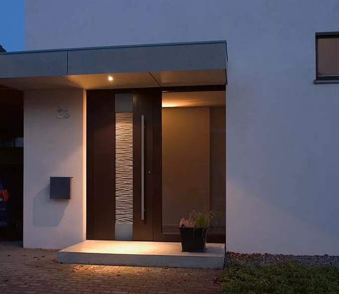 Nowoczesne drzwi zewnętrzne z ciemnym skrzydłem, pionowym podświetlonym przeszkleniem o fakturze i długim pochwytem, pod zadaszeniem przy wejściu do domu.