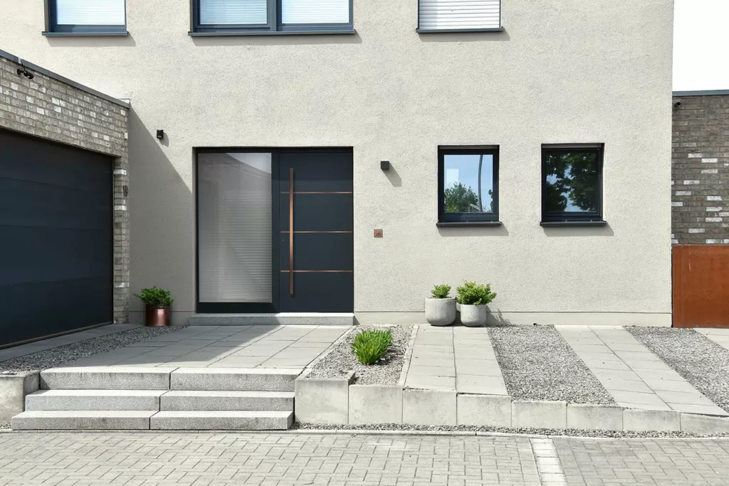Nowoczesne antracytowe drzwi zewnętrzne z bocznym naświetlem, długim pionowym uchwytem i miedzianymi liniami, na tle minimalistycznej elewacji domu z betonowymi płytami, żwirem i donicami.
