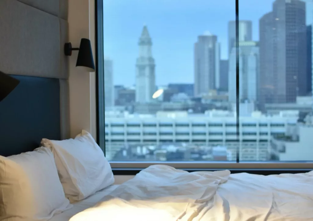 Łóżko z białą, pogniecioną pościelą w nowoczesnym pokoju, stojące przy dużym oknie z szybą dźwiękoszczelną z widokiem na panoramę miejskich wieżowców za dnia.