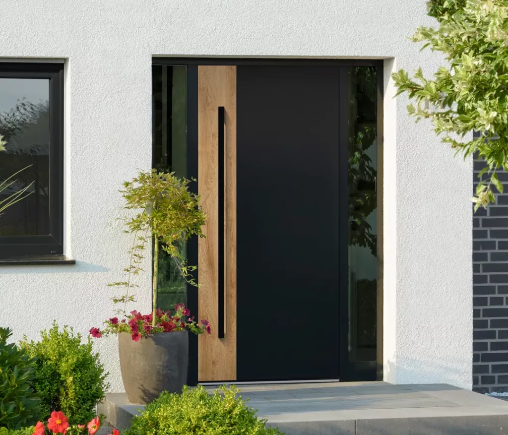 Nowoczesne drzwi zewnętrzne w kolorze antracytowym z pionowym drewnianym panelem i długim uchwytem, z wąskimi naświetlami po bokach, w białej elewacji i otoczeniu zadbanego ogrodu z kwiatami.