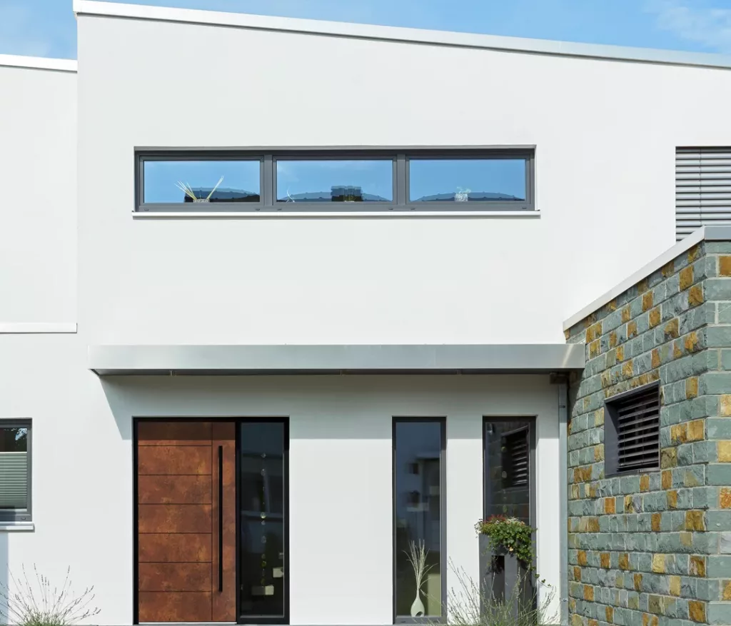 Nowoczesne drzwi zewnętrzne w kolorze rdzawej stali z długim pionowym uchwytem i wąskim naświetlem bocznym, osadzone w białej elewacji domu z fragmentem kamiennej okładziny i żwirową ścieżką przed wejściem.