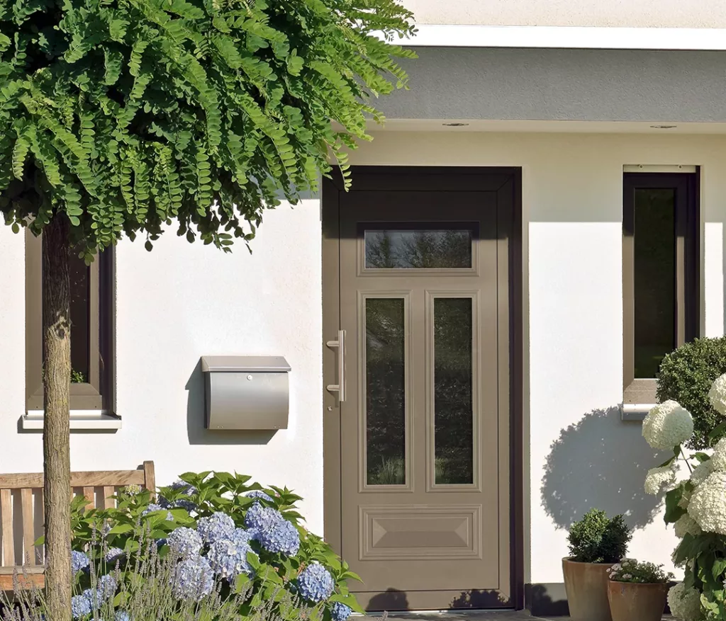 Jasnobrązowe drzwi wejściowe z trzema przeszkleniami (dwoma wąskimi i jednym u góry) w białej elewacji domu, obok srebrnej skrzynki na listy, ławki i kwitnących hortensji.