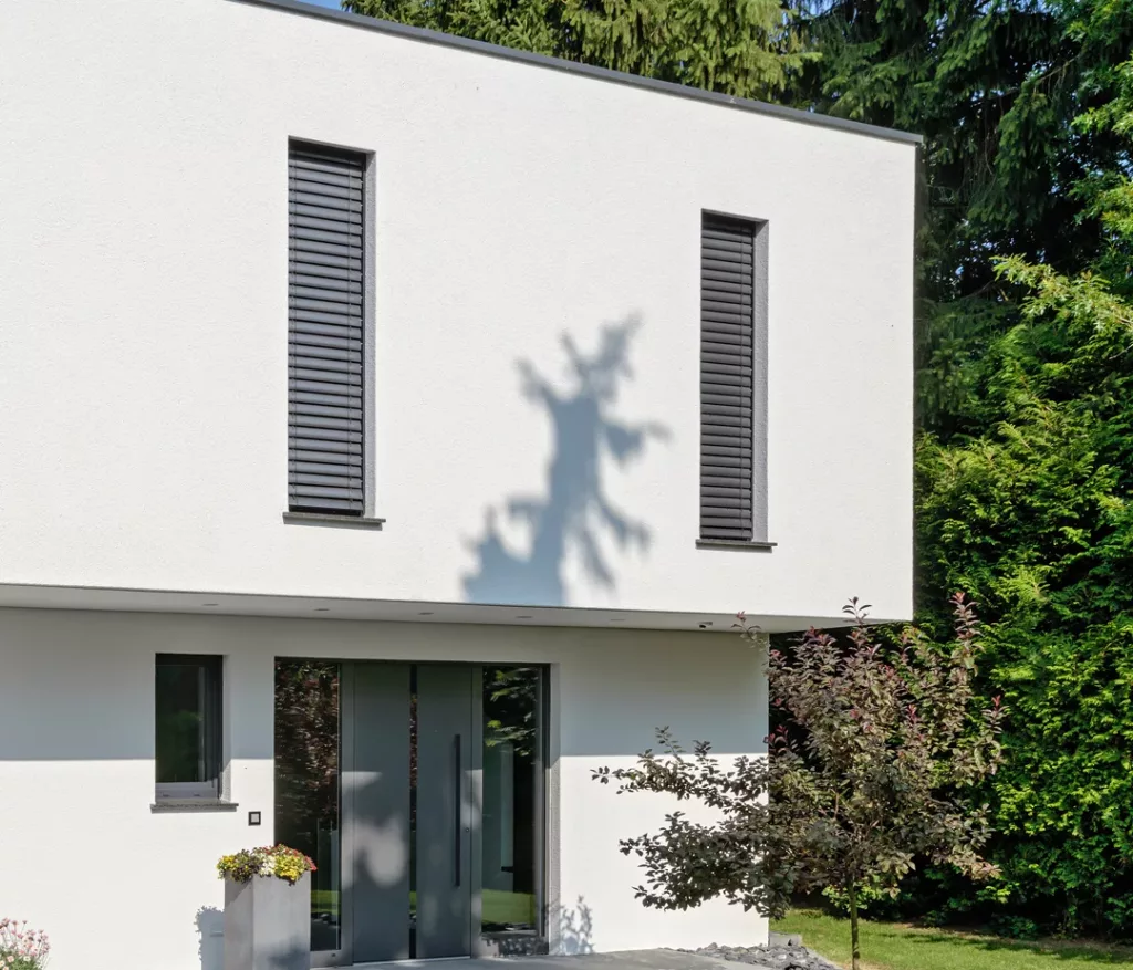 Nowoczesne, grafitowe drzwi zewnętrzne dwuskrzydłowe z wysokim pionowym uchwytem i wąskimi przeszkleniami, w białej minimalistycznej elewacji domu, otoczone płytowym podejściem i rabatami z ciemnym grysem.