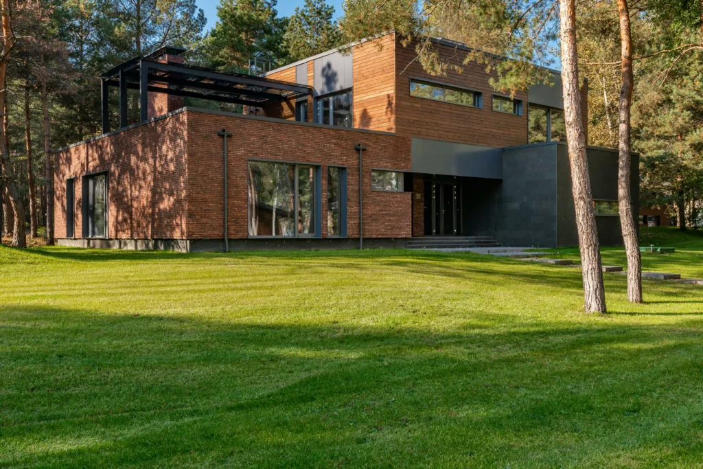 Nowoczesny, dwukondygnacyjny dom o płaskim dachu z ceglaną i drewnianą elewacją, dużymi oknami aluminiowymi i czarną pergolą na tarasie, stojący na zadbanym trawniku wśród sosnowego lasu.