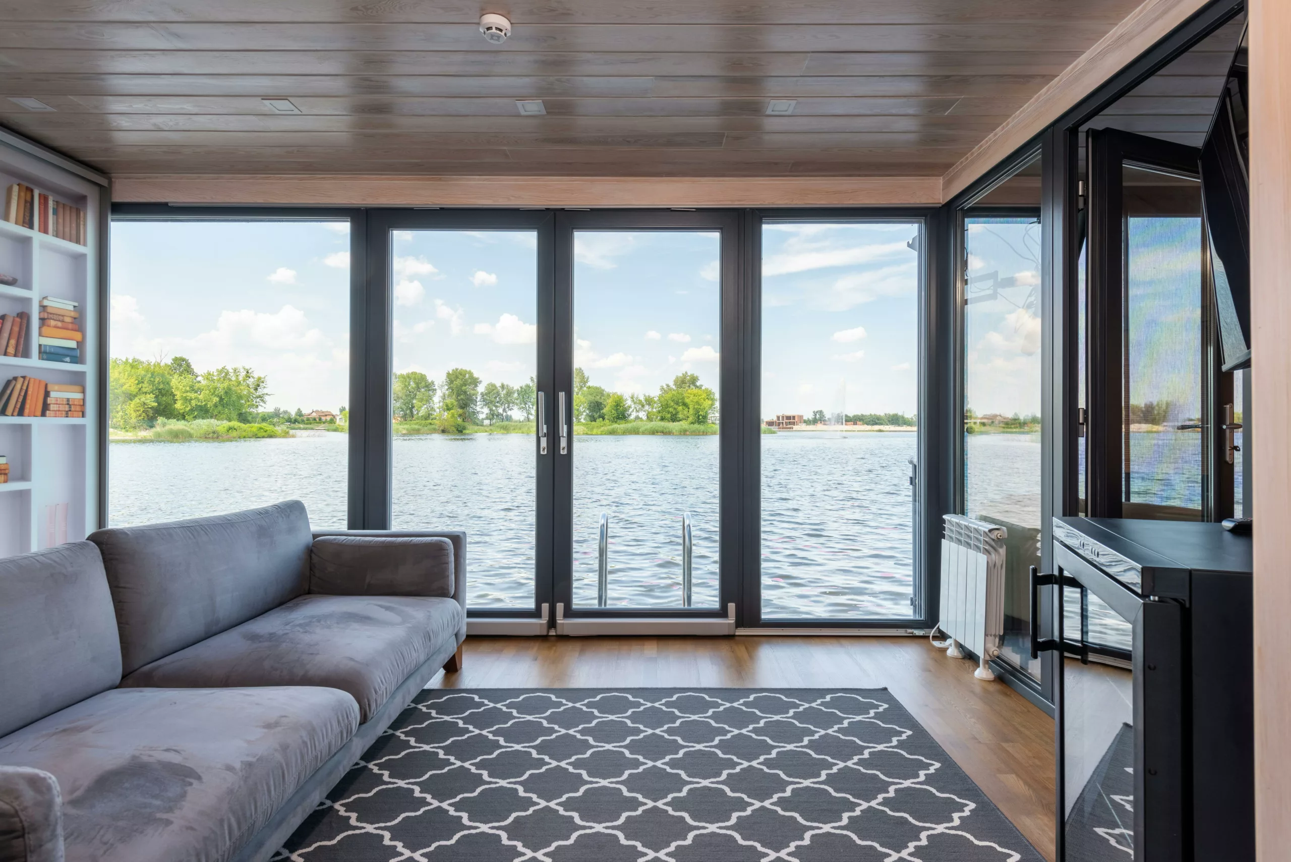 Salon z panoramicznymi drzwiami PSK na jezioro, drabinką do wody; szara kanapa i dywan w romby na drewnianej podłodze.
